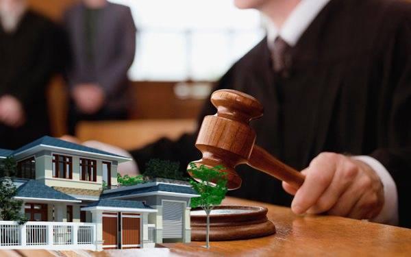 một người đang ngồi cầm búa thẩm phán, bên cạnh là hình ảnh ngôi nhà