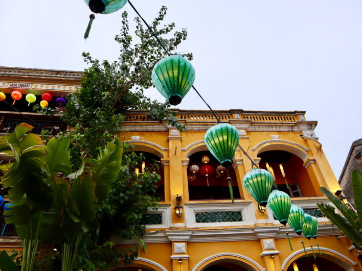 Mặt trước ngôi nhà màu vàng tại Hội An, Việt Nam, nhiều đèn lồng, cây cối nhiệt đới.