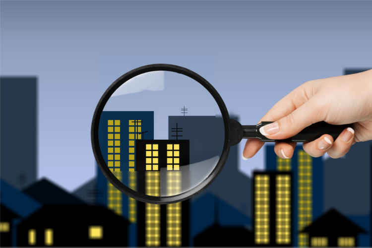 Ảnh thiết kế bàn tay cầm một chiếc kính lúp, nền phía sau là mô hình dãy nhà ở