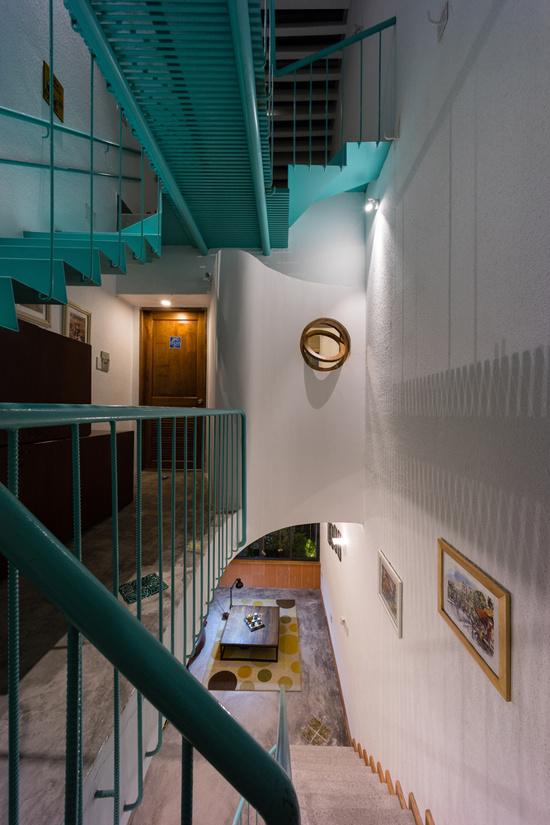 Ảnh chụp từ trên xuống một cầu thang nhà ống có lan can màu xanh, tranh treo tường, bên dưới là bàn trà phòng khách