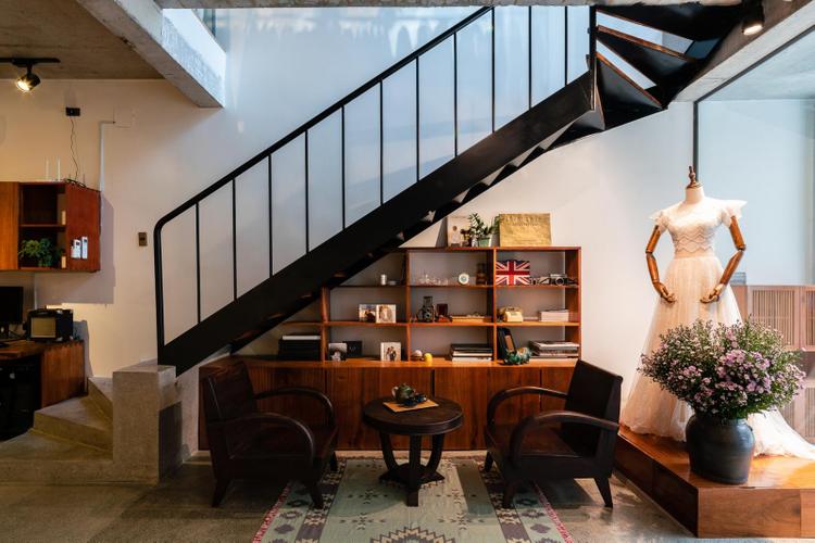 Cầu thang màu đen, bên dưới là phòng khách với kệ đồ, bàn ghế gỗ, manocanh mặc váy trắng, lọ hoa...