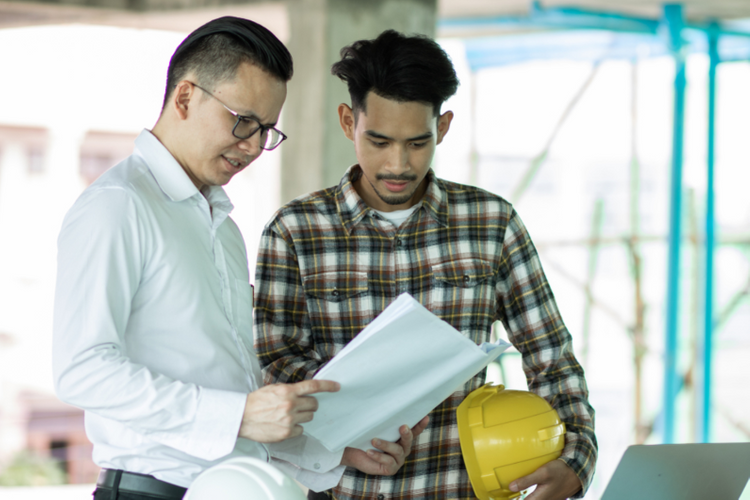 Hai người đàn ông đứng trao đổi với nhau, người mặc sơ mi trắng đeo kính cầm giấy tờ, người mặc áo kẻ caro cầm mũ bảo hộ lao động.