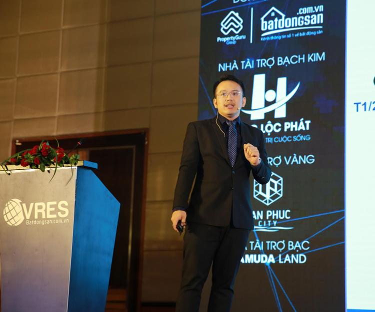 Chân dung một người đàn ông trẻ tuổi, mặc vest đen, đeo kính trắng đứng trên bục sân khấu.