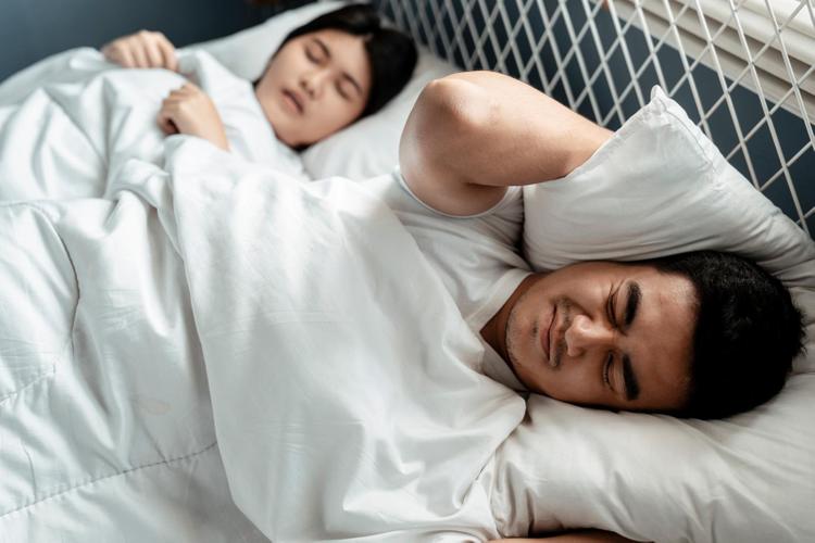 người đàn ông cau có, khó chịu  vì tiếng ồn khi đang nằm ngủ cạnh vợ