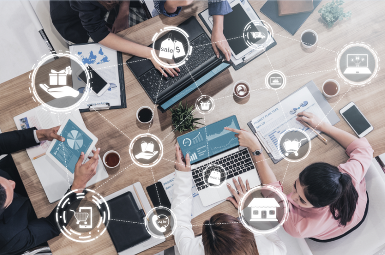 5 người ngồi xung quanh chiếc bàn để thảo luận với nhau, trên bàn có điện thoại và máy tính