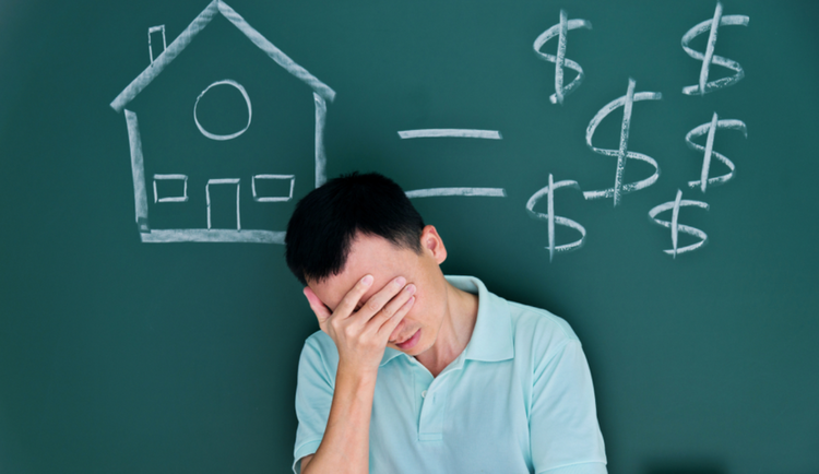 Ảnh người đàn ông cúi đầu tay ôm trán, phía sau lưng là tấm bảng vẽ hình ngôi nhà và $