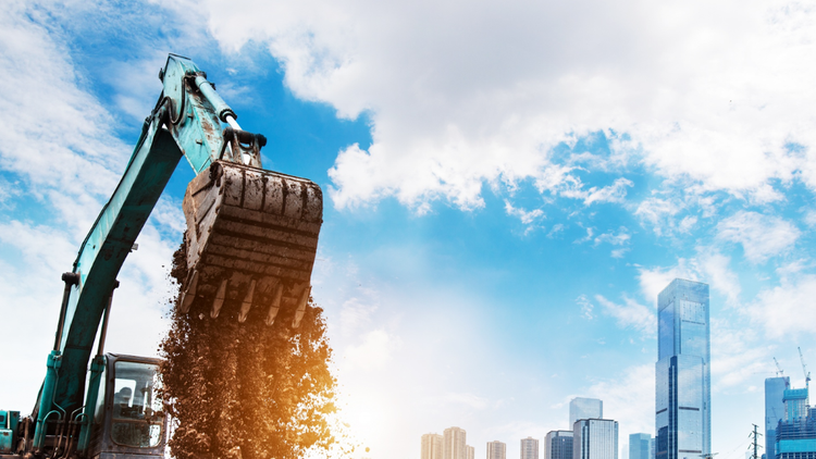 An excavator with a whole bucket of dirt is silhouetted against a bright blue sky
