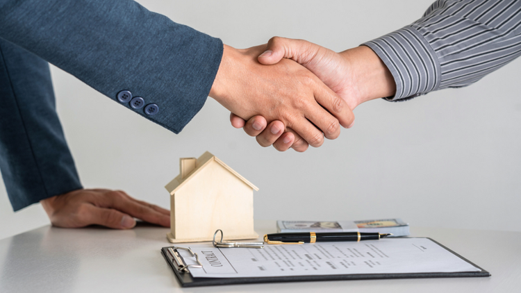 Two people shake hands over a desk with a small wooden house model 
