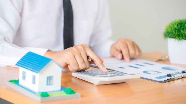 A person in business attire uses a calculator at a desk with property documents and a small model house in the foreground.