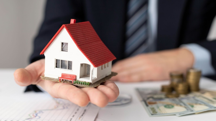 A man carrying a model house in his palm with money and documents placed on the table