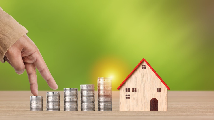 Hand moving across stacked coins towards a small wooden house, symbolising saving for a home.
