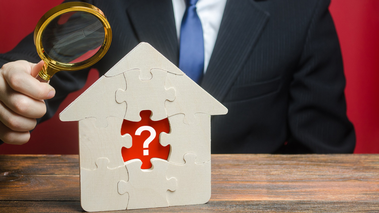 A person holds a magnifying glass on a wooden puzzle house with a question mark