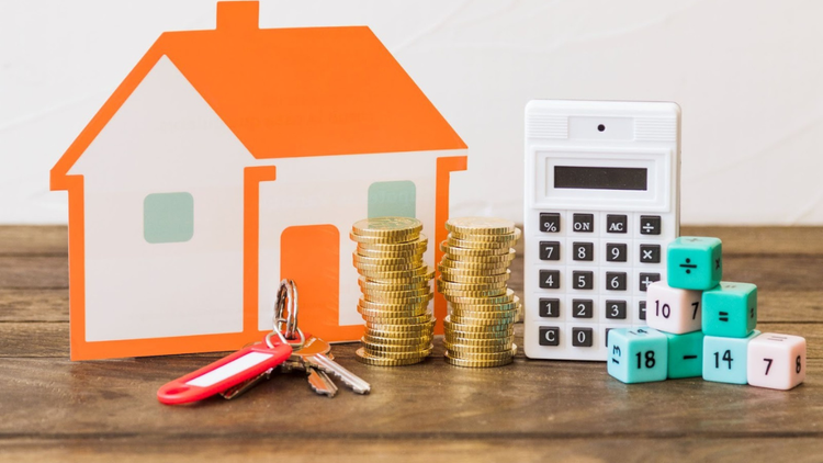 A paper house, keys, stacked coins, a calculator and math blocks are placed on a wooden table