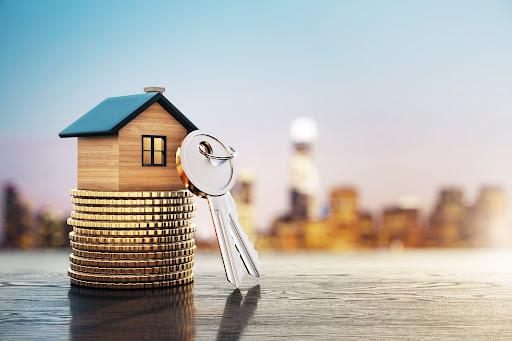 A miniature wooden house placed on stacks of coins with house keys beside it, set against a blurred city skyline.