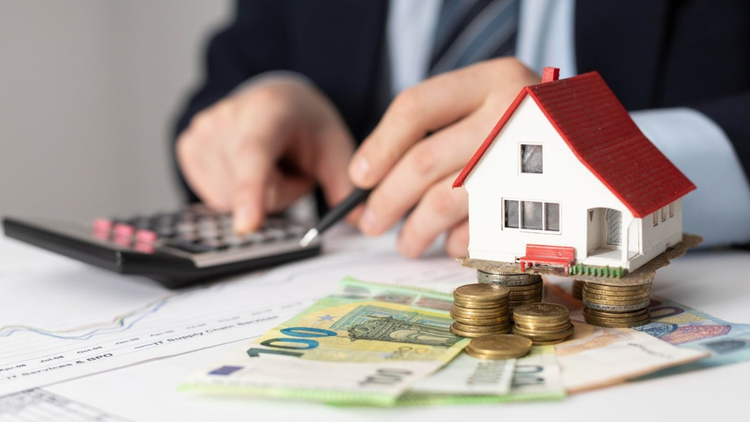 A man's hand holds a calculator and coins on a desk, next to a small model house, suggesting financial planning.