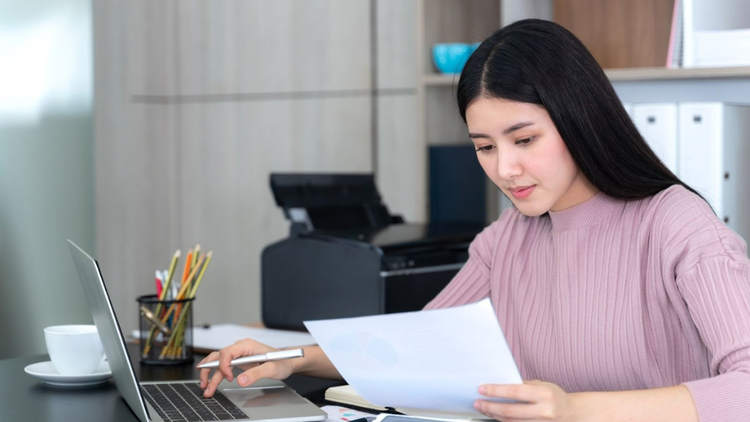 A young woman typing on her laptop and going through a document at the same time