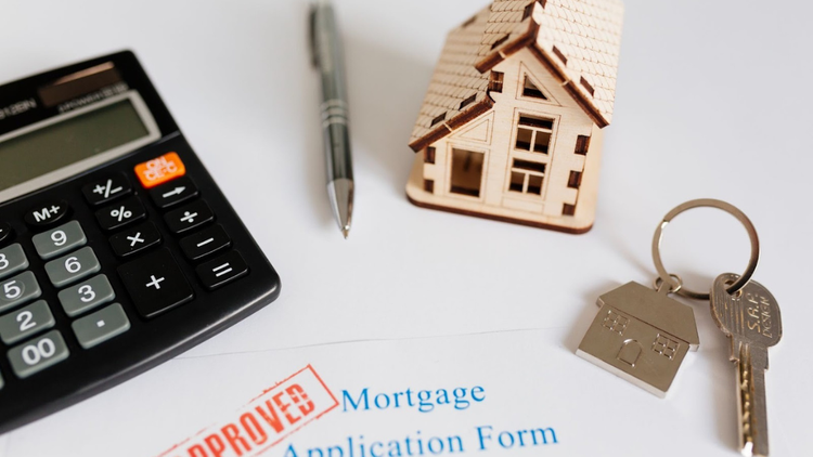 A calculator, pen, and small wooden house model sit on a table alongside a "Mortgage Application Form" stamped "Approved" with a house key.