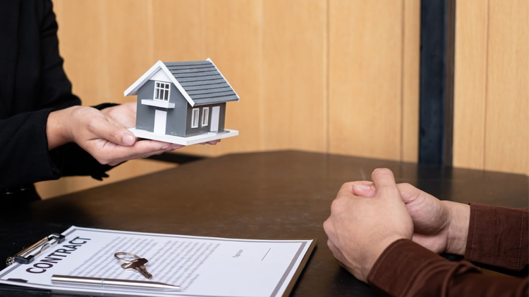 Person presenting a small model house across a table with a contract and keys.