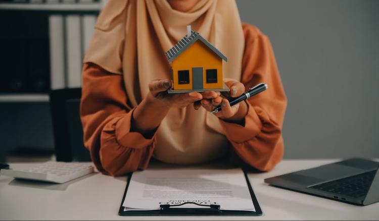 Close-up of a woman in a hijab holding a house model and pen while signing documents.