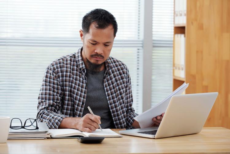 A casually dressed man working in an office, writing in a notebook and holding documents
