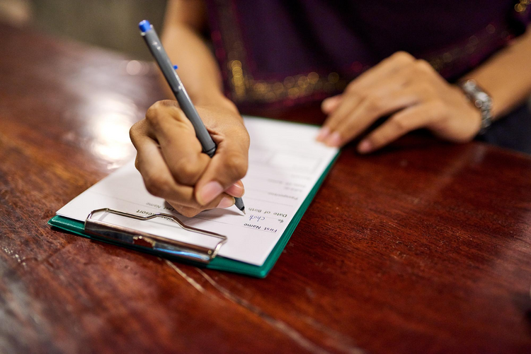 A person writes on a form secured to a clipboard on a dark wooden table