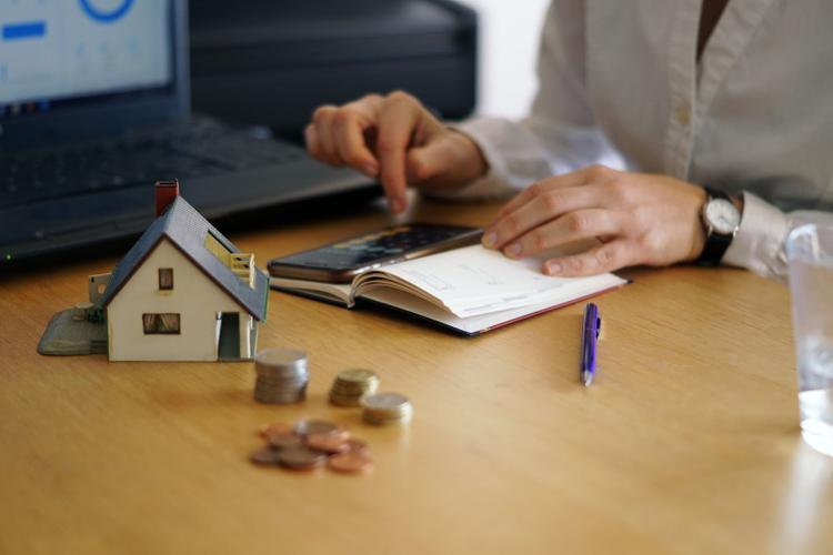 Person calculating home expenses with a model house, coins, and a notebook on a desk.