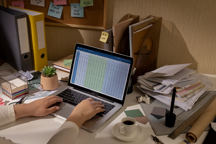 Overhead view of a messy desk with hands typing on a laptop displaying a large spreadsheet (Excel/data analysis).
