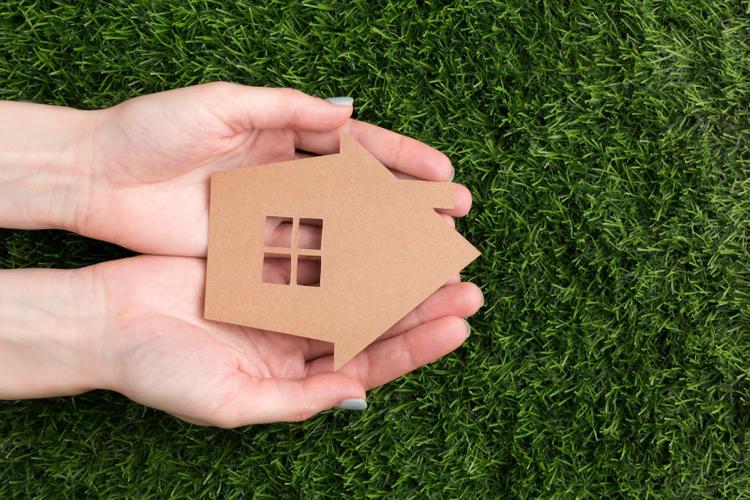 Close-up of hands gently holding a small cardboard house on a grassy surface.