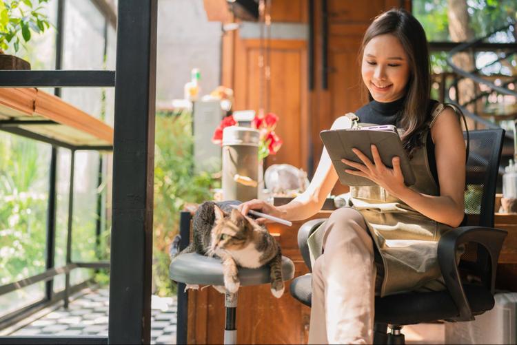 A Malaysian woman working on her tablet while gently patting her cat sitting on a stool beside her.